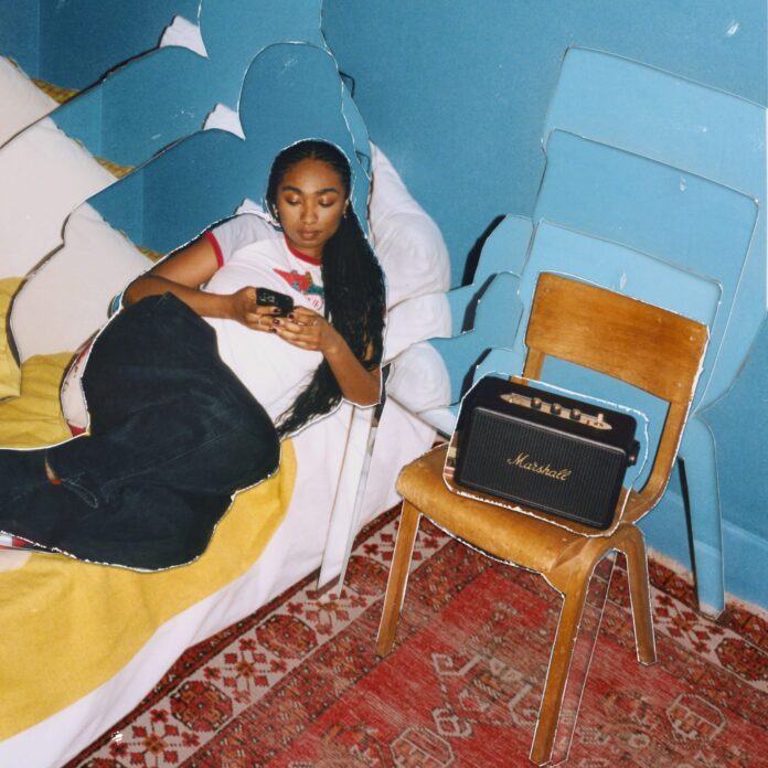 ผู้หญิงอยู่บนเตียงในห้องที่ตกแต่งอย่างอบอุ่นและมีสีสัน ข้างเตียงมีลำโพง Marshall Kilburn III วางอยู่บนเก้าไม้อย่างโดดเด่น