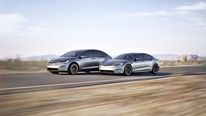 Tesla Model S และ Model X วิ่งเคียงข้างกันบนถนนในทะเลทราย ภายใต้แสงแดดยามเช้า ฉากหลังเป็นภูเขาและท้องฟ้าโปร่ง