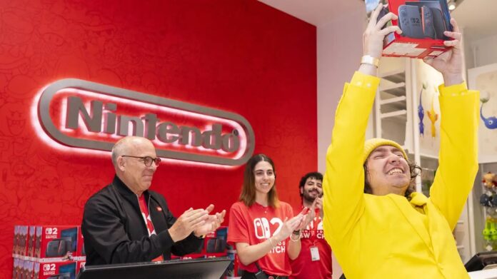 ชายสวมชุดสีเหลืองชูกล่อง Nintendo Switch 2 ด้วยความดีใจในร้าน Nintendo ขณะพนักงานรอบข้างปรบมือแสดงความยินดี