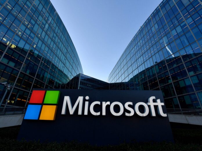 ภาพป้ายโลโก้ Microsoft ขนาดใหญ่ตั้งอยู่หน้าสำนักงานที่มีอาคารกระจกสูงสองฝั่งล้อมรอบ