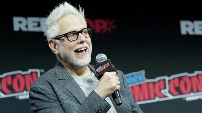 James Gunn สวมแว่นและสูทลายสก็อตกำลังถือไมโครโฟนพูดบนเวทีในงาน New York Comic Con