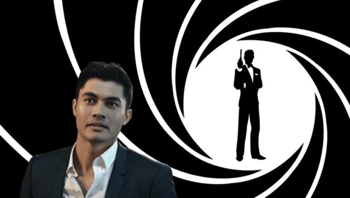 Henry Golding ในชุดสูทยืนหน้าฉากกราฟิกโลโก้ปืนของแฟรนไชส์ James Bond