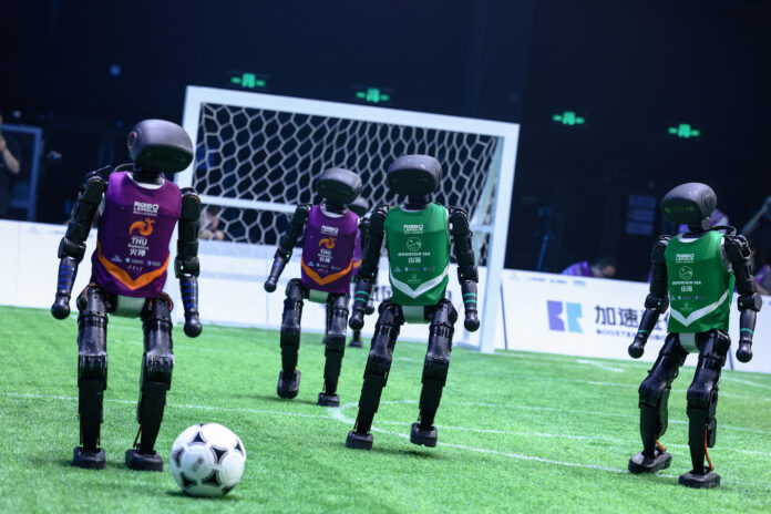 ภาพหุ่นยนต์ AI รูปร่างมนุษย์สวมเสื้อทีมสีม่วง (THU Robotics) และสีเขียว (Mountain Sea) กำลังแข่งขันฟุตบอลบนสนามหญ้าเทียม