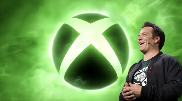 Phil Spencer ยืนยิ้มอยู่ด้านหน้าโลโก้ Xbox ขนาดใหญ่ที่เปล่งแสงสีเขียว
