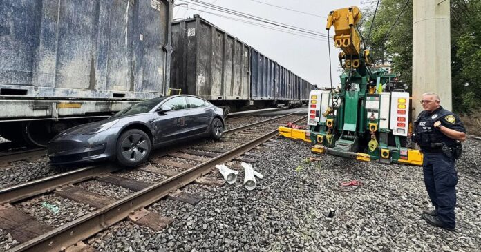 รถยนต์ Tesla สีดำจอดค้างอยู่บนรางรถไฟขนส่งสินค้า โดยมีรถลากกำลังเตรียมเคลื่อนย้าย และเจ้าหน้าที่ตำรวจยืนอยู่ใกล้ ๆ