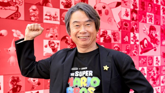 ชิเงรุ มิยาโมโตะ สวมเสื้อ The Super Mario Bros. Movie ยิ้มและชูแขนท่ามกลางฉากหลังที่มีตัวละครจากเกม Nintendo