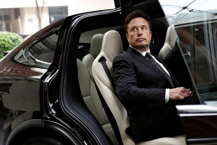Elon Musk สวมสูทสีดำและเนคไทลายจุดนั่งอยู่ในรถ Tesla