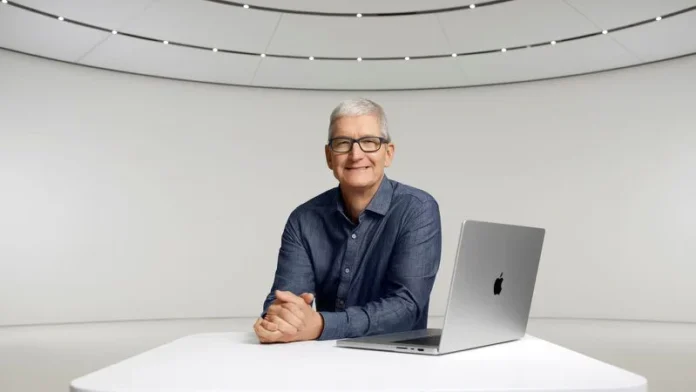 Tim Cook นั่งอยู่ที่โต๊ะสีขาวในห้องประชุมดีไซน์ล้ำ พร้อม MacBook วางอยู่ด้านหน้า สวมเสื้อเชิ้ตแขนยาวสีกรมท่า และยิ้มอย่างเป็นกันเอง