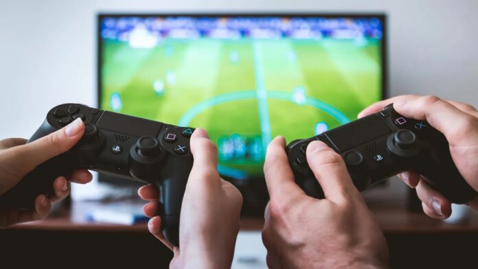 มือสองคู่กำลังถือจอยคอนโทรลเลอร์ PS4 เล่นเกมฟุตบอลบนหน้าจอโทรทัศน์