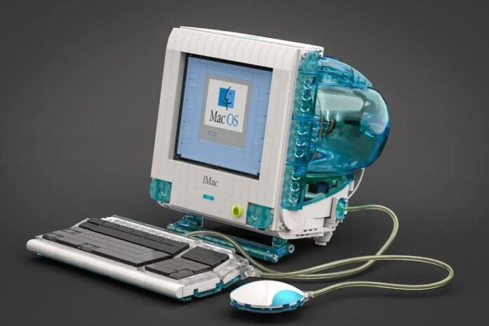 โมเดล LEGO จำลองคอมพิวเตอร์ iMac G3 สี Bondi Blue แบบ all-in-one พร้อมคีย์บอร์ดและเมาส์ “ฮอกกี้พัค” ที่เชื่อมต่อด้วยสายแบบใส ภาพแสดงดีไซน์โปร่งใสด้านหลังเครื่องและหน้าจอที่แสดงโลโก้ Mac OS บนพื้นหลังสีเทา วางอยู่บนพื้นสีเทาเข้ม