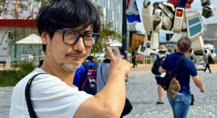 Kojima ในลุคใส่แว่นยกมือชี้ไปที่หุ่นกันดั้มขนาดใหญ่กลางแจ้ง