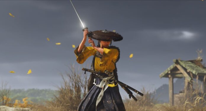 Atsu จาก Ghost of Yōtei ในชุดเสื้อเหลืองชักดาบพร้อมฟันกลางทุ่งหญ้า โดยมีใบไม้ร่วงและศาลาไม้เก่าด้านหลัง