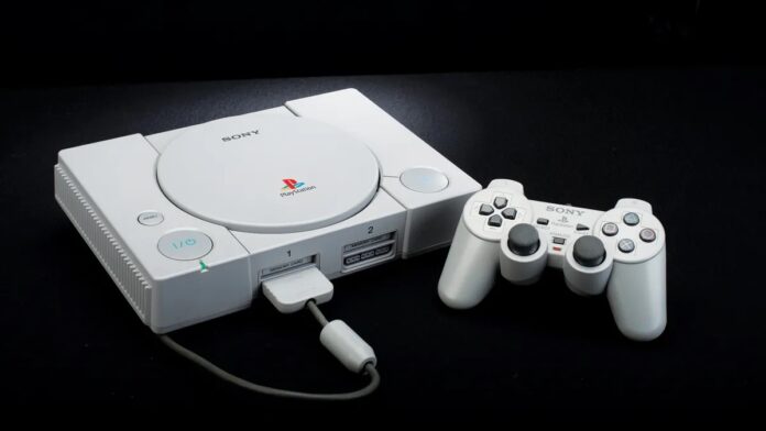 เครื่องเล่นเกม Sony PlayStation รุ่นแรกสีเทาวางอยู่บนพื้นสีดำ พร้อมจอยคอนโทรลเลอร์ DualShock วางอยู่ข้างๆ