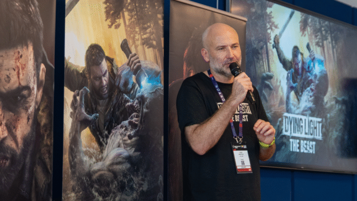 ภาพ Tymon Smektala ขณะพูดในงานแถลงข้อมูลเกม Dying Light: The Beast โดยมีโปสเตอร์โปรโมตตัวเกมอยู่ด้านหลัง