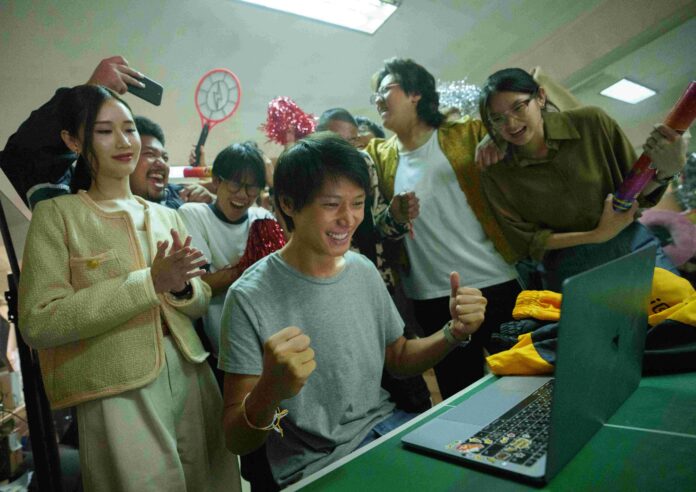 ภาพจากซีรีส์ Netflix “สงคราม ส่งด่วน” ตัวเอกนั่งหน้าคอมพิวเตอร์ แสดงท่าดีใจ ขณะที่เพื่อน ๆ รอบตัวส่งเสียงเชียร์และปรบมือร่วมแสดงความยินดี