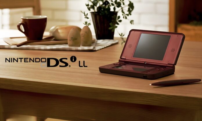 เครื่องเล่นเกมพกพา Nintendo DSi LL สีแดงเข้ม วางอยู่บนโต๊ะไม้ข้างสไตลัส พร้อมถ้วยกาแฟ ขนมปัง และต้นไม้ในฉากหลัง บรรยากาศอบอุ่นแบบในบ้าน