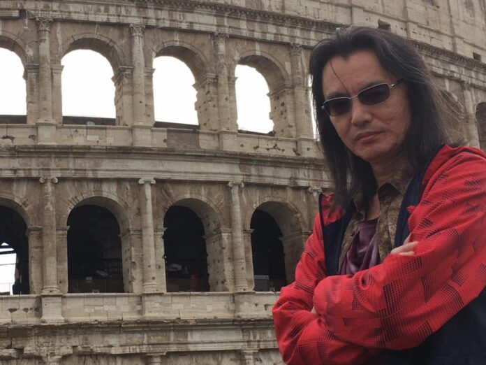 ภาพ Tomonobu Itagaki สวมแว่นกันแดดและเสื้อแจ็กเก็ตสีแดงยืนกอดอกหน้าโคลอสเซียมในกรุงโรม ประเทศอิตาลี