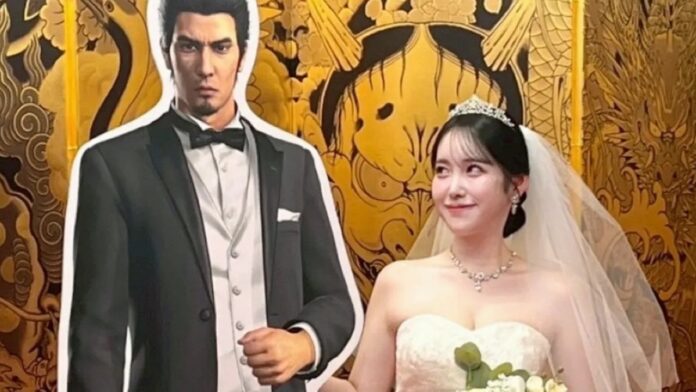 หญิงสาวชาวญี่ปุ่นสวมชุดเจ้าสาวยืนคู่กับแผ่นคัตเอาต์ของ Kazuma Kiryu จากซีรีส์ Like a Dragon ภายในงานอีเวนต์ฉลองครบรอบ 20 ปีของแฟรนไชส์