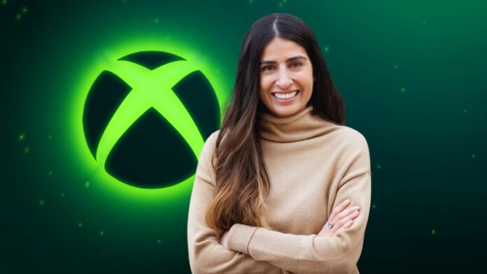 Asha Sharma ยืนยิ้มกอดอกอยู่หน้าพื้นหลังโลโก้ Xbox สีเขียวเรืองแสง