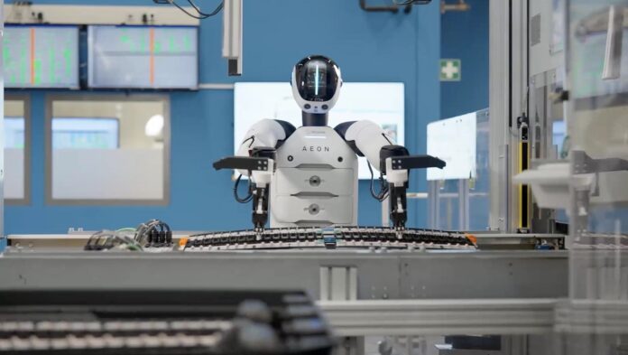 หุ่นยนต์ฮิวแมนนอยด์สีขาวกำลังยืนทำงานบนสายการผลิตภายในโรงงานอุตสาหกรรม พร้อมแขนกลและอุปกรณ์เครื่องจักรรอบด้าน