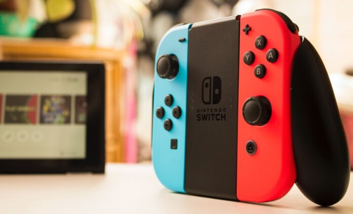 Joy-Con สีแดงและน้ำเงิน วางบนโต๊ะ โดยมีเครื่อง Switch อยู่ด้านหลัง