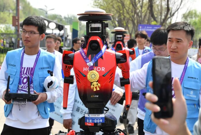 หุ่นยนต์ฮิวแมนนอยด์สีแดงแบรนด์ HONOR สวมเหรียญรางวัล ยืนอยู่ท่ามกลางผู้คนระหว่างงานวิ่งมาราธอนกลางแจ้ง