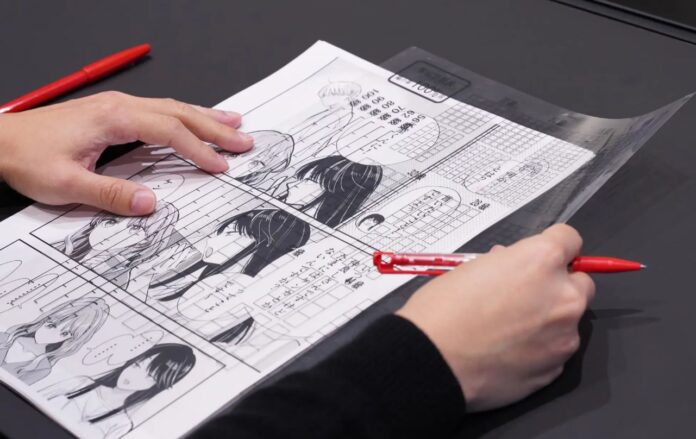 มือของบุคคลกำลังวาดและแก้ไขหน้ามังงะบนกระดาษ โดยใช้ปากกาสีแดง