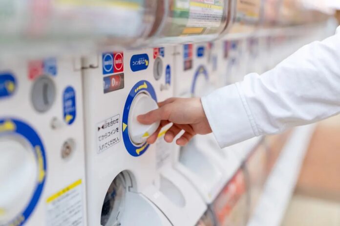 มือของบุคคลกำลังสุ่มตู้กาชาปอง (ตู้สุ่มของเล่น) ที่เรียงต่อกันหลายเครื่องในร้านค้า