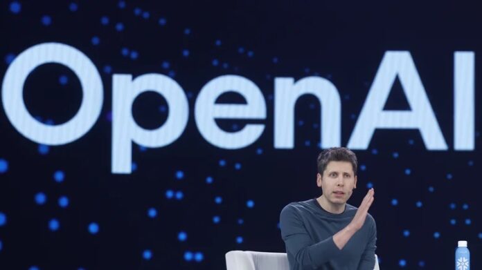 โลโก้ OpenAI บนจอขนาดใหญ่ พร้อม Sam Altman นั่งอยู่บนเวทีด้านหน้า