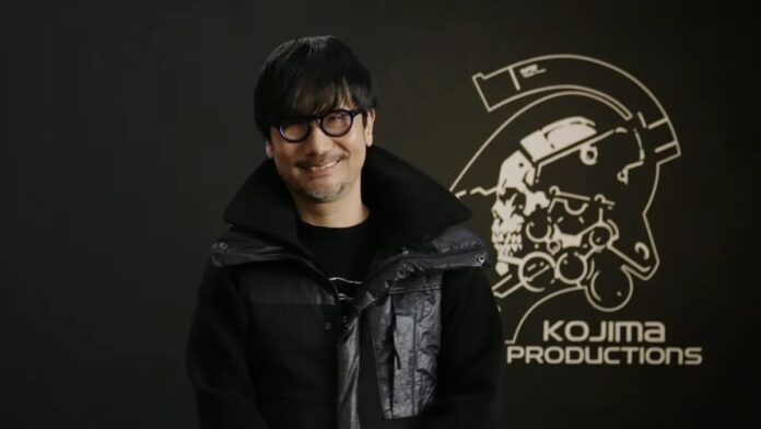 Hideo Kojima ในชุดเสื้อแจ็กเก็ตสีดำ ยืนอยู่หน้าโลโก้สตูดิโอ Kojima Productions