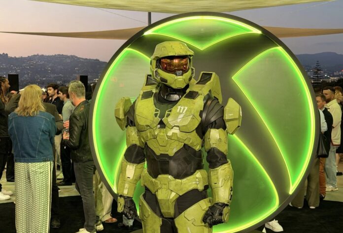 หุ่น Master Chief เกราะเขียว ยืนโพสต์ท่าหน้าโลโก้ Xbox สีเขียวขนาดใหญ่