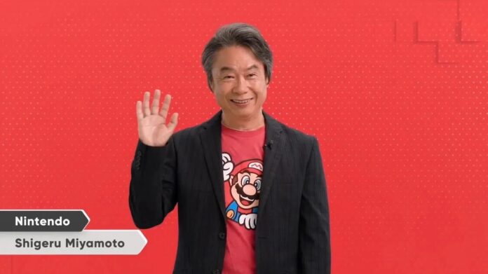 ชิเงรุ มิยาโมโตะ ผู้สร้าง Mario และ Zelda สวมเสื้อลายมาริโอ้ โบกมือทักทายหน้าฉากหลังสีแดง