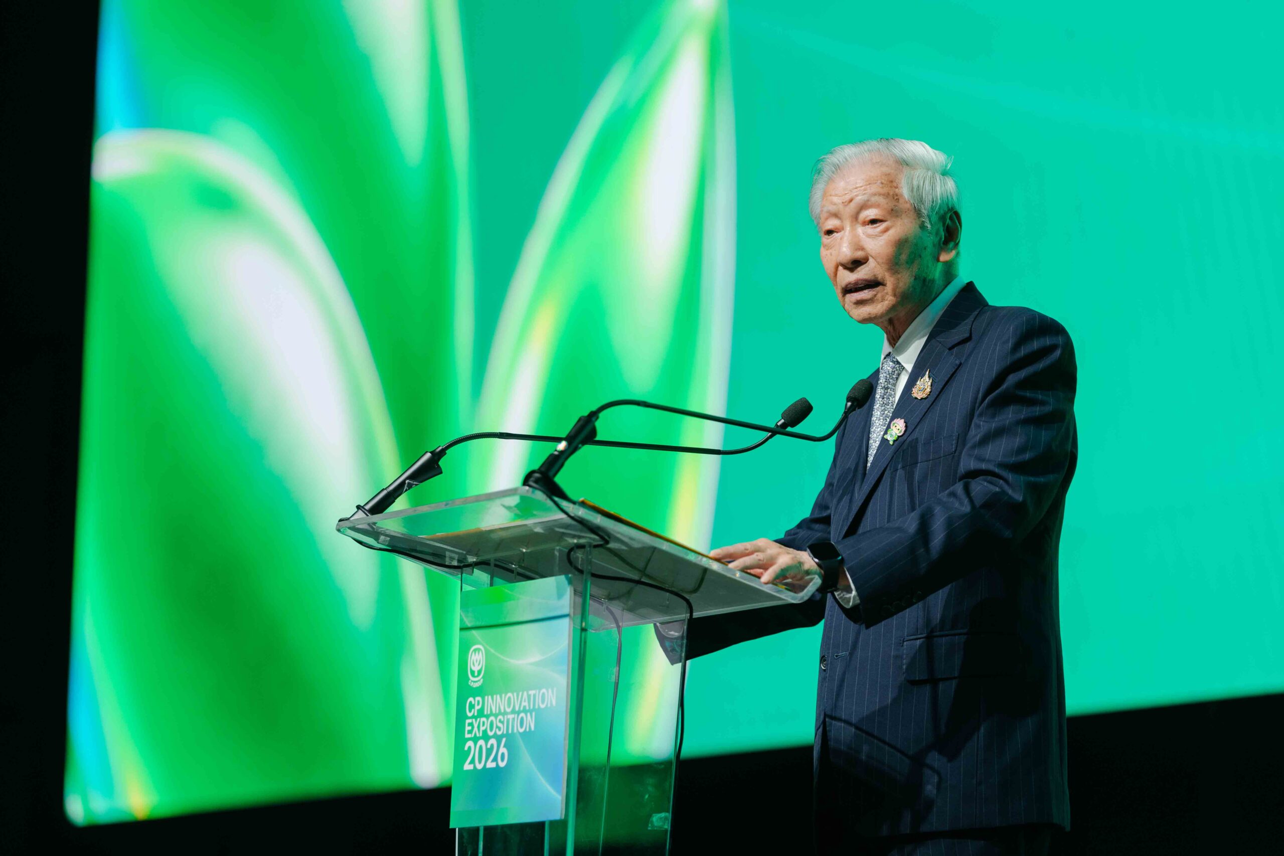 ดร.อาชว์ เตาลานนท์กล่าวที่โพเดียมใสหน้าฉากสีเขียวและป้ายชื่องาน CP Innovation Exposition 2026